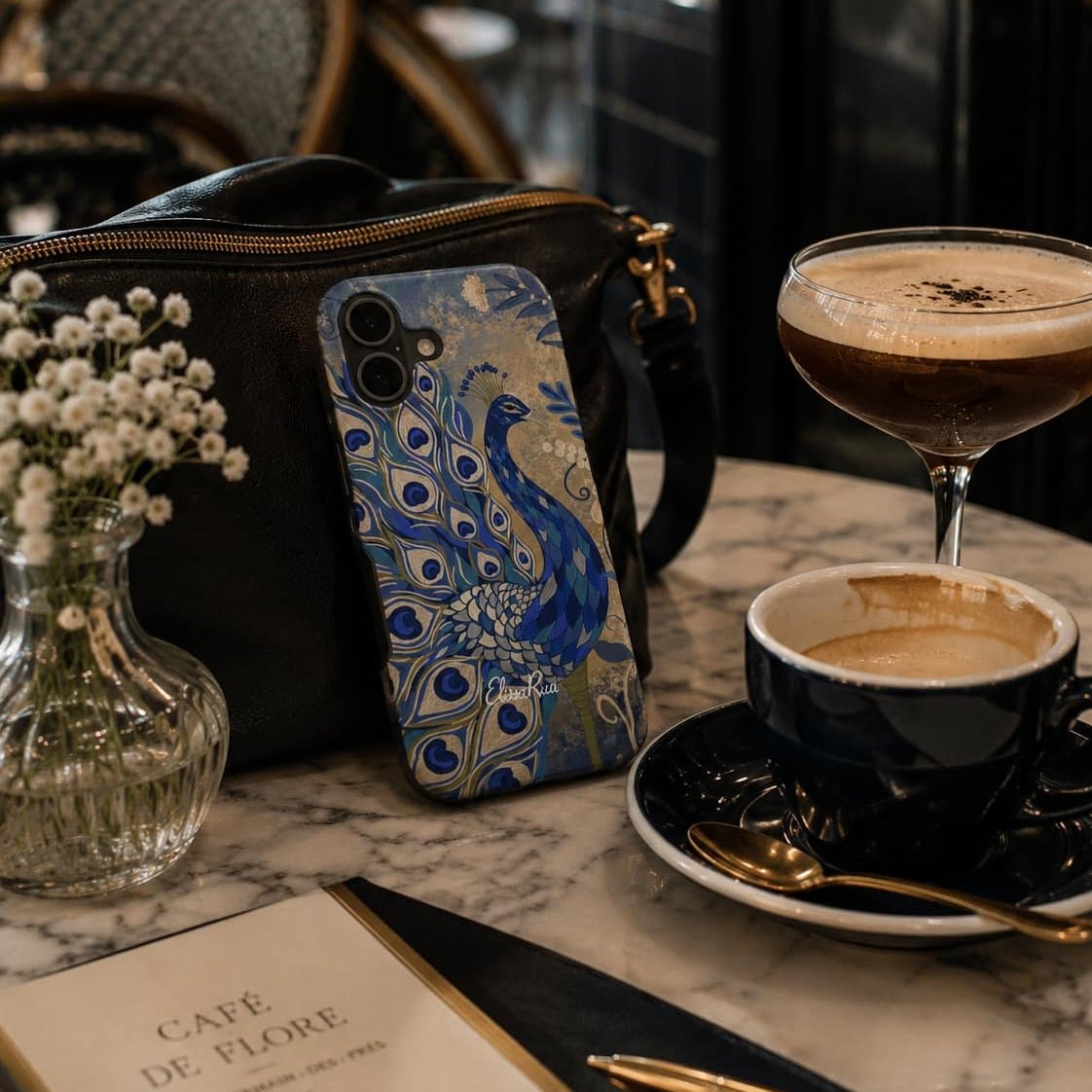 peacock-patterned phone case, coffee cup, and cocktail on a marble table.