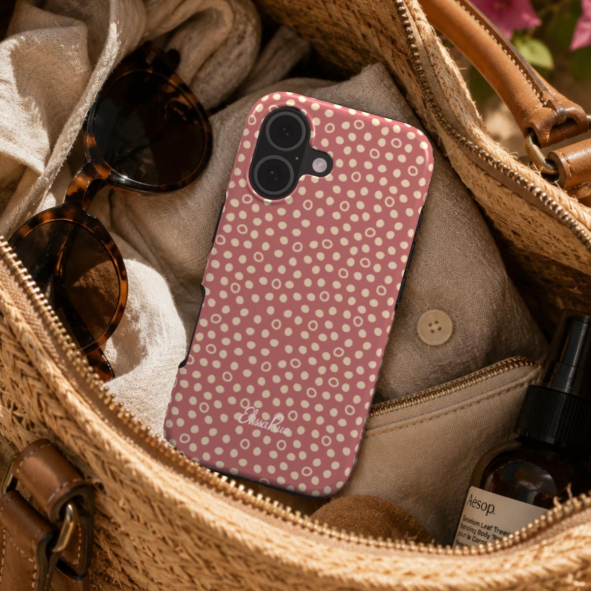 Phone case with dusty pink polka dot pattern in an open beige handbag with sunglasses and a bottle.
